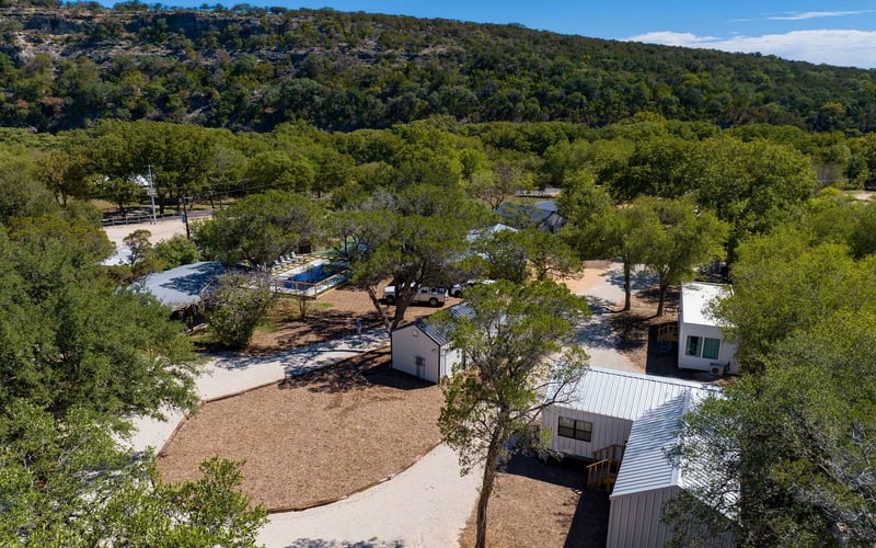 Scenic view of a countryside New Braunfels Vacation Rental property with modern cabins, gravel pathways, shaded trees, and a tennis court, set against a hillside backdrop.