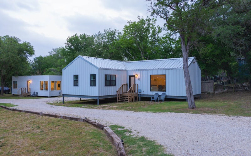 Exterior view of modern white metal-clad tiny houses with small decks, set on a gravel road in a wooded area during evening.