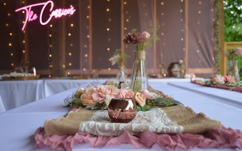 Decorated wedding reception table with pink and peach flowers, candles, and burlap runners, in a rustic outdoor setting with string lights and a neon sign.