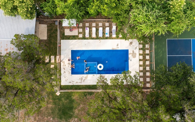 An aerial view of a swimming pool with five children playing in it, surrounded by trees, lounge chairs, and a tennis court.