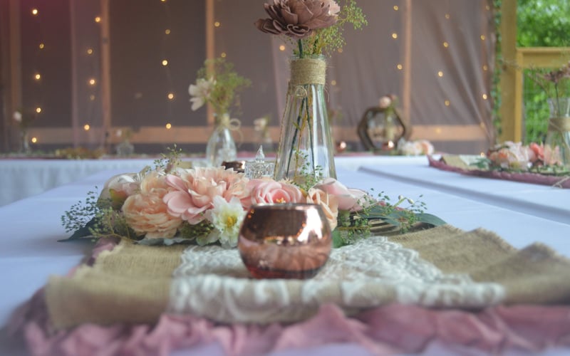 A wedding scape table decorated with pink and white flowers, a rose gold candle holder, glass vases with flowers, and rustic fabric accents in an outdoor setting with warm fairy lights.