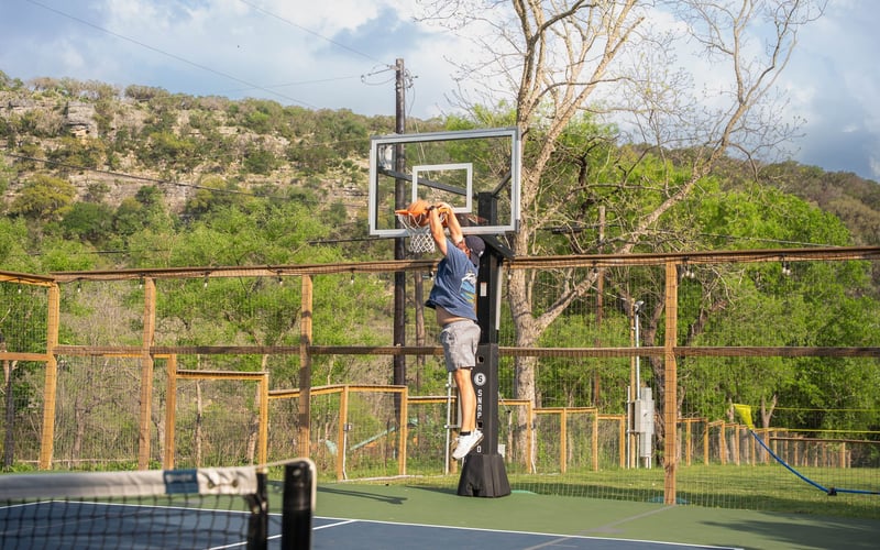 A person hanging from a basketball hoop on an outdoor court, about to dunk the ball, with wooden fencing, trees, and hills in the background.