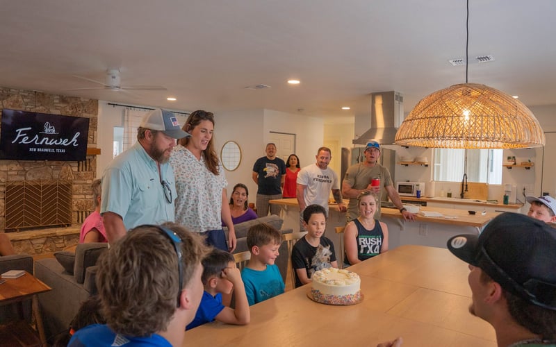 People gathered around a birthday cake in a cozy kitchen, with children sitting at a table and adults standing behind, celebrating inside a home in New Braunfels, Texas.