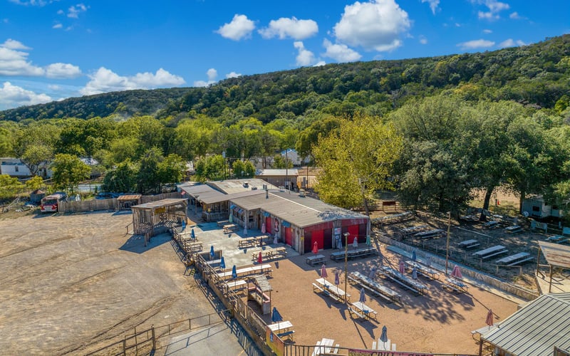 Aerial view of an outdoor rustic restaurant and bar with picnic tables, umbrellas, and a 'Grill is Open' sign, set against a scenic hillside and lush greenery.