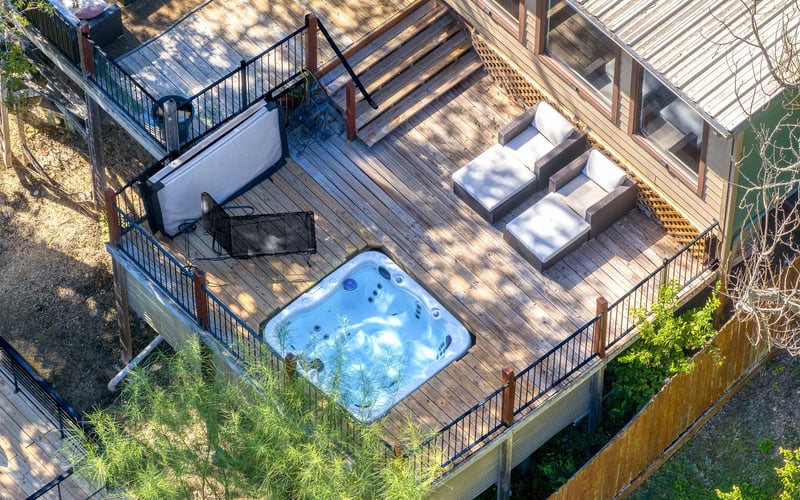 Private wooden deck with a built-in hot tub, lounge chairs, and black metal railing, attached to a New Braunfels Vacation Rental surrounded by greenery.