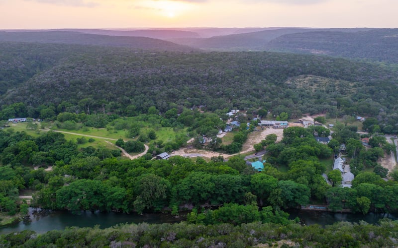 Aerial view of a lush green landscape with a river running through the foreground, a small community with houses and buildings, and rolling hills or mountains in the background during sunset.