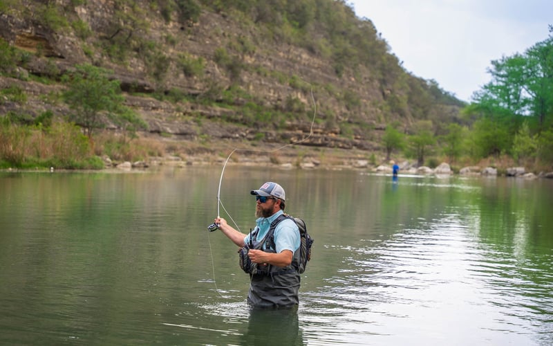 A man fly fishing in a river with a mountainous landscape in the background. He's wearing a baseball cap, sunglasses, and fishing vest, standing waist-deep in the water, holding a fishing rod.