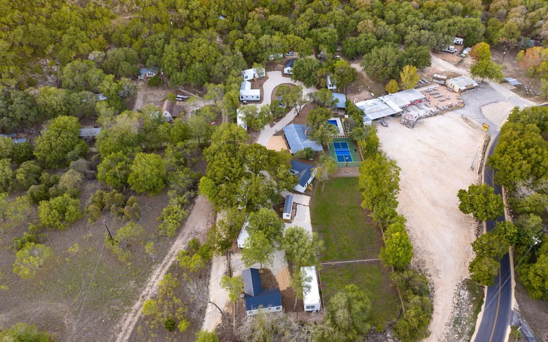 Aerial view of a New Braunfels Vacation rental with modern cottages, a tennis court, and lush greenery, nestled among rolling hills near a winding road.