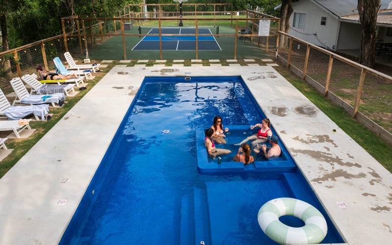 Resort pool with hot tub. There are five people sitting in the hot tub, and one person lying on a lounge chair on the poolside. Seven lounge chairs and fenced-in tennis or pickleball court in background