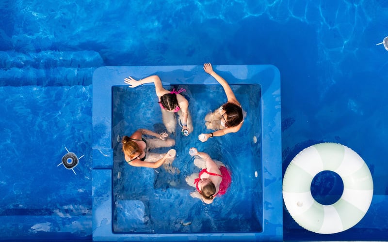 Four women and a girl in swimsuits enjoying a pool party, raising glasses in a toast inside a swimming pool, with a life preserver floating nearby.