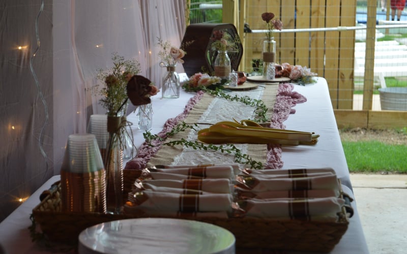 Decorated outdoor table set for a celebration with floral arrangements, cloth napkins, cutlery, and plates in a tented area with string lights and a wooden fence.