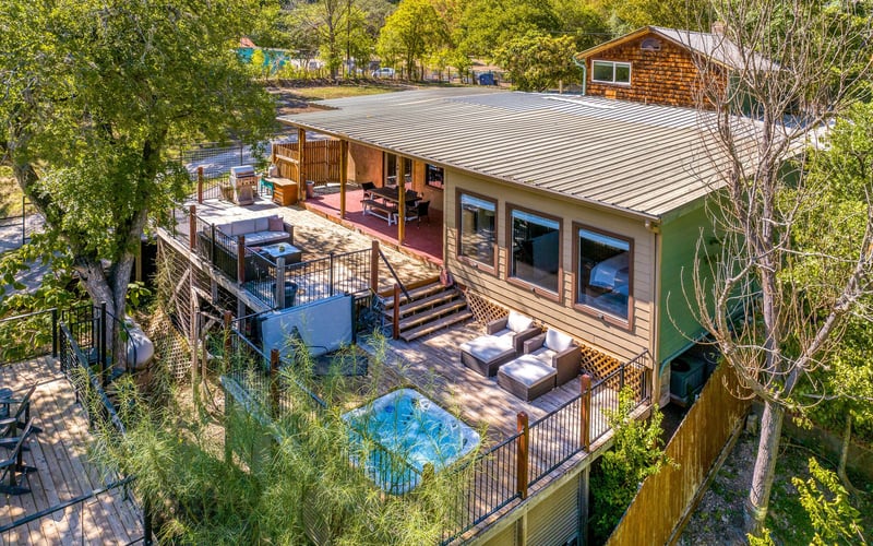 Aerial view of a spacious New Braunfels Vacation Rental with a covered patio, outdoor lounge seating, a hot tub, and multiple decks, surrounded by lush greenery.