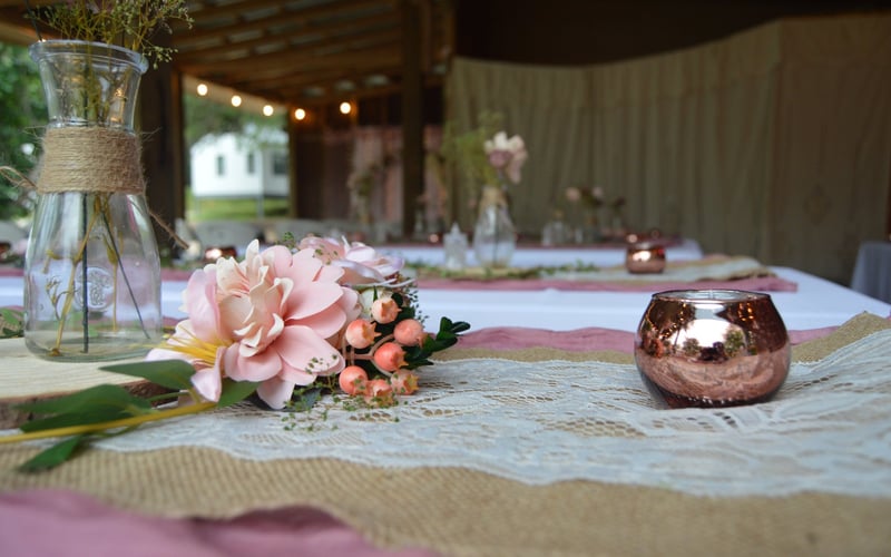A decorated table with a pink and beige floral centerpiece, a pink glass candle holder, and vases with flowers, set in a rustic outdoor setting with string lights.