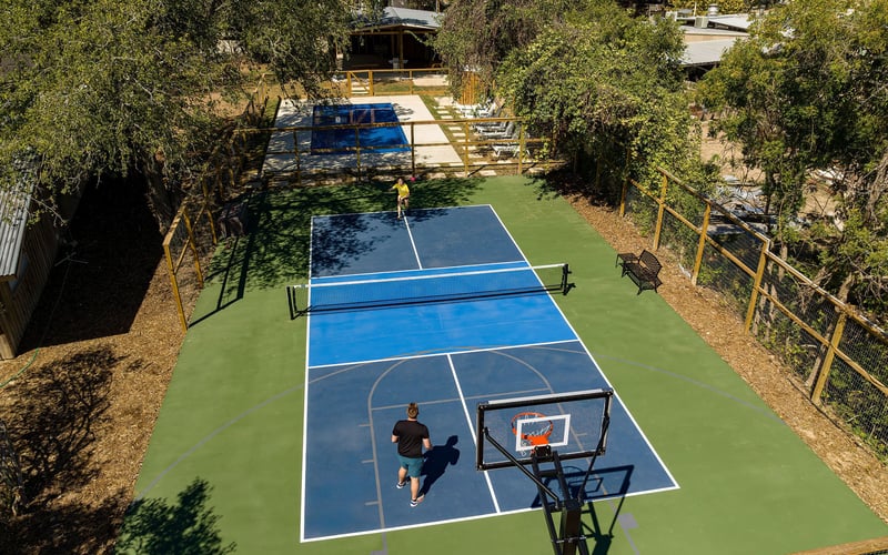 Multi-purpose outdoor sports court surrounded by trees and fencing. The court is painted green and blue, featuring a basketball hoop in the foreground and a pickleball net in the middle. Two people are engaged in a game, one near the net and the othe