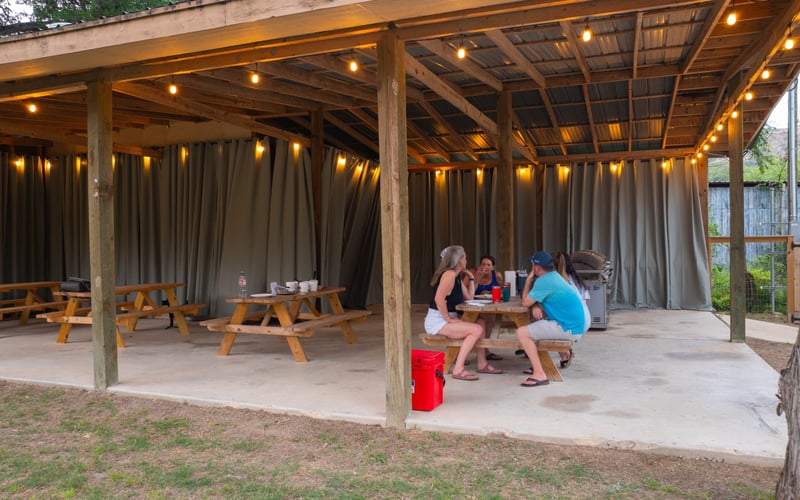 Group of four people sitting at a picnic table under a wooden pavilion with string lights, in a backyard setting with fencing and trees.