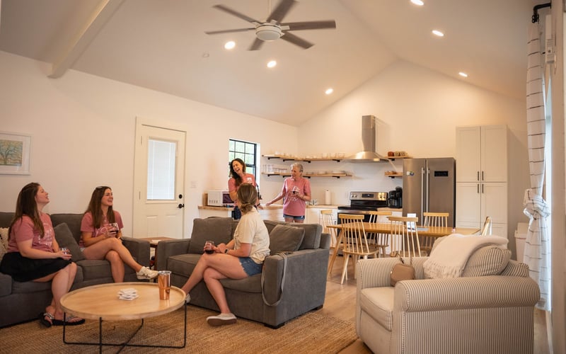 Women in pink T-shirts socialize in a living room with a kitchen in the background, some sitting on sofas and an armchair, holding drinks, others standing and chatting, with a ceiling fan and recessed lighting.