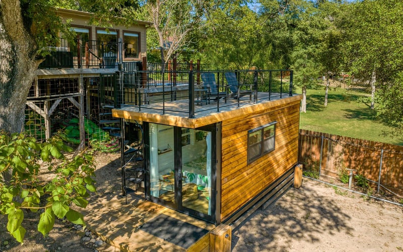 Modern tiny home with a rooftop deck, black metal railing, glass doors, and a spiral staircase, nestled in a wooded setting at a New Braunfels Vacation Rental.