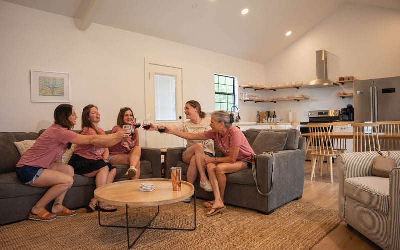 Five women sitting on a gray couch in a living room, smiling and toasting glasses of red wine, with a kitchen in the background.