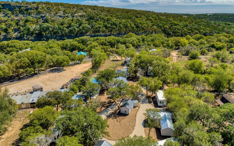 Scenic countryside New Braunfels Vacation Rental with modern cabins, shaded trees, winding pathways, and a pickleball court, set against a lush hillside backdrop.