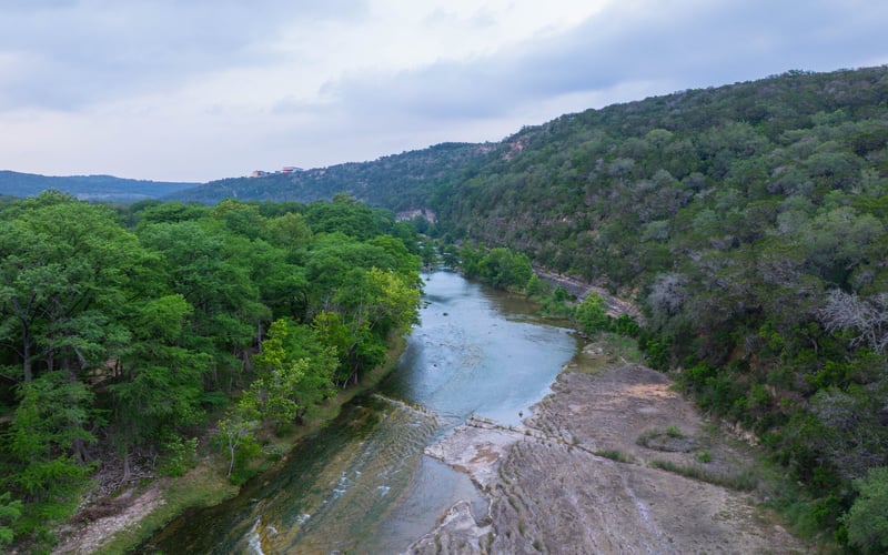 A scenic view of a river flowing through a lush green valley with forested hills on either side, under a partly cloudy sky.