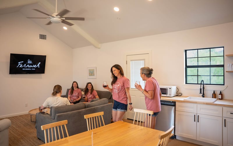Five women gathering and chatting in a cozy living room with a kitchenette, some sitting on a sofa and some standing, all holding glasses of wine, during daytime with natural light coming through the window.