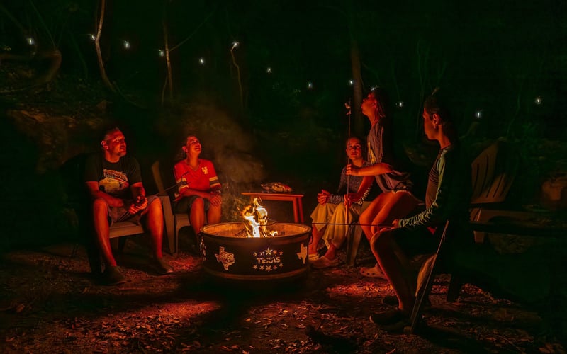 Five people sitting around a campfire at night, with a dark forest background and fireflies flying above.